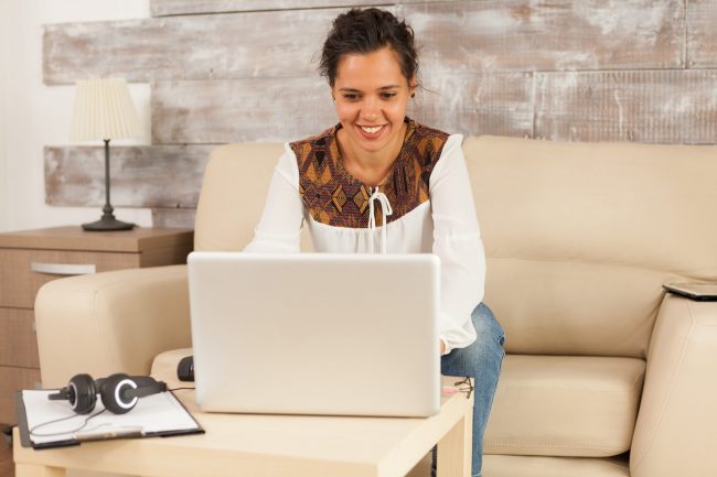 Photo of woman in living room with laptop illustrates blog: "20 Remote Job Interview Questions You Should be Ready to Answer"