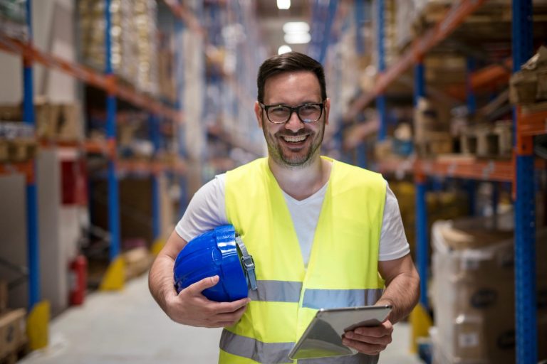 Photo of man smiling in warehouse with safety best and helmet under his arm illustrates blog: "How To Become a Warehouse Supervisor"