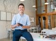 Photo of smiling young man sitting on a desk illustrates blog: "Office Assistant vs Office Manager: What Is the Difference?"