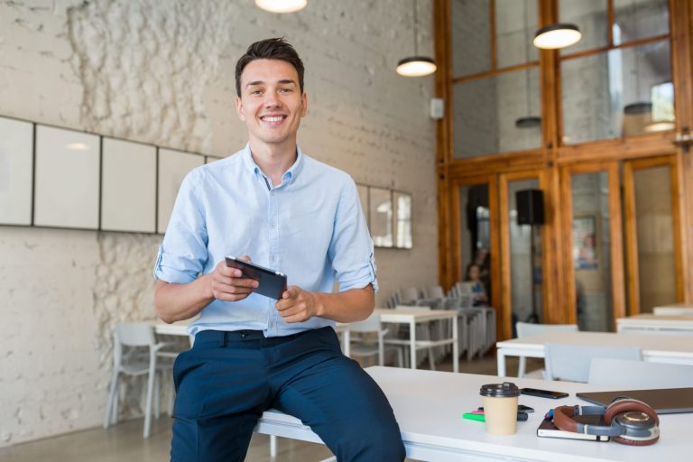 Photo of smiling young man sitting on a desk illustrates blog: "Office Assistant vs Office Manager: What Is the Difference?"